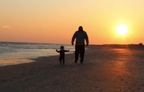 A silhouette of a man and a child walking on a beach at sunset, with the sun setting over the ocean.