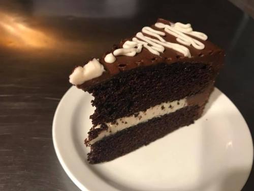 A slice of chocolate cake with a creamy filling and white icing drizzled on top, served on a white plate.