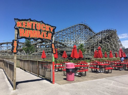 Sign for "Kentucky Rumbler" roller coaster with red umbrellas and picnic tables in front of the wooden track.