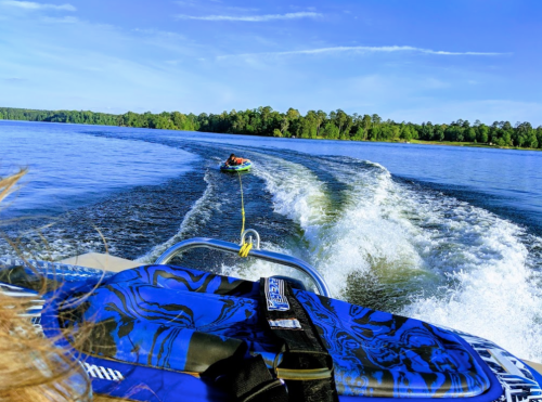 A person is being towed on a tube behind a boat on a calm lake, surrounded by trees and a clear blue sky.