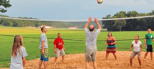 A group of people playing beach volleyball on a sandy court with a net, surrounded by green fields and trees.