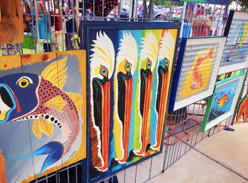 Colorful artwork displayed at an outdoor market, featuring fish and pelicans in vibrant styles.