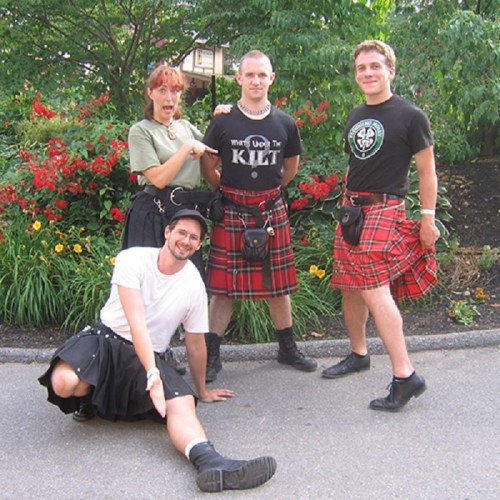 Four people in kilts pose outdoors, surrounded by colorful flowers, with one person sitting and three standing.