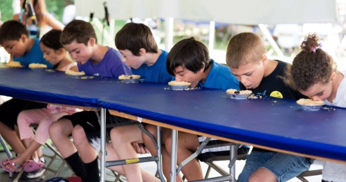 A group of children compete in a pie-eating contest, leaning over tables with pies in front of them.