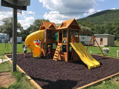 Children playing on a colorful wooden playground with slides and a climbing structure, set against a green hillside.