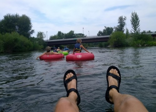 A person in sandals relaxes while floating on a river, with others in colorful inner tubes nearby and greenery in the background.