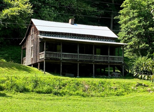 A rustic wooden house with a metal roof, surrounded by greenery and a grassy area.