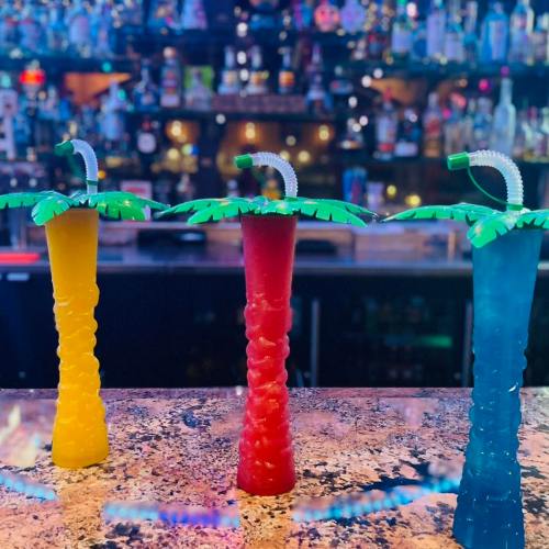 Three colorful tropical drinks with palm tree straws sit on a bar, with a blurred background of bottles.