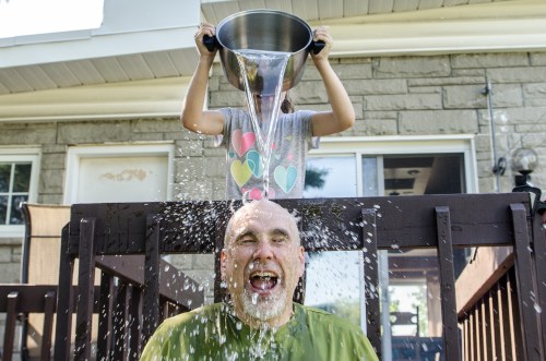 A child pours water from a large pot over a laughing adult's head, creating a splash in a backyard setting.