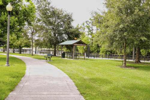 A paved path leads to a gazebo by a calm river, surrounded by green grass and trees.
