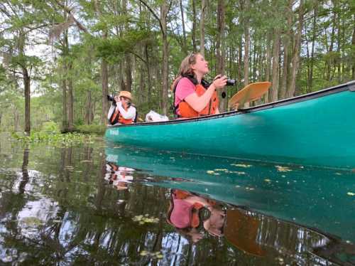 Two people in orange life jackets kayak through a serene, wooded area, capturing photos amidst the calm water.