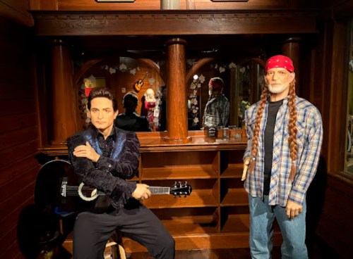 Two lifelike wax figures in a wooden setting: one with a guitar and the other in a plaid shirt and bandana.