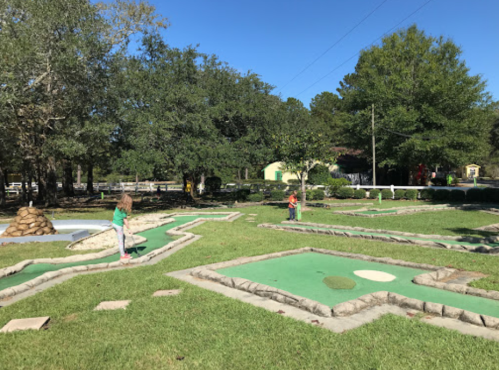 Two children play mini-golf on a sunny day, surrounded by green grass and trees.