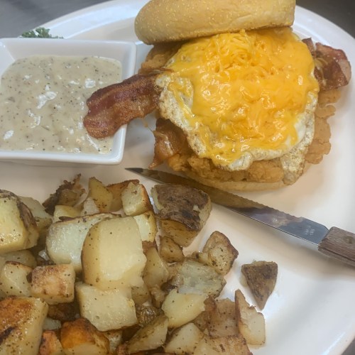 A plate with a crispy chicken sandwich topped with cheese and bacon, served with a side of potatoes and gravy.