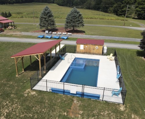 Aerial view of a fenced swimming pool with a shed and shaded seating area, surrounded by green fields and trees.