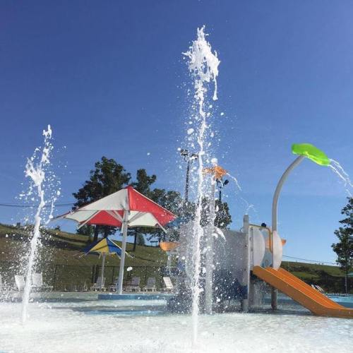 A sunny pool scene featuring water fountains, a colorful slide, and shaded umbrellas.