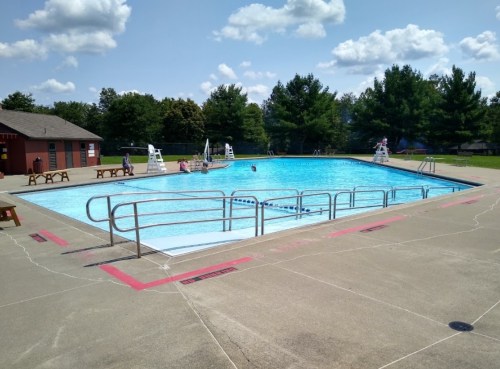 A sunny outdoor swimming pool with lifeguard chairs, surrounded by trees and a concrete deck.