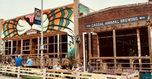 Colorful mural on a building with outdoor seating at a brewery, people enjoying drinks on a sunny day.