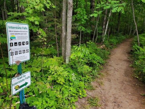 A trail sign directing hikers to Interstate Falls, surrounded by lush green foliage and a dirt path.