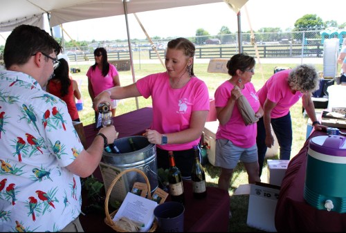 A woman in a pink shirt pours wine for a customer at an outdoor event, with others in pink shirts assisting nearby.