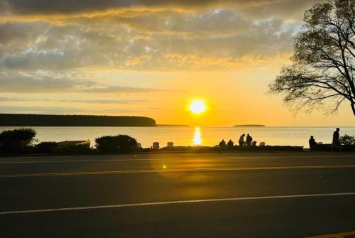 Sunset over a calm lake, with silhouettes of people sitting by the shore and trees lining the background.