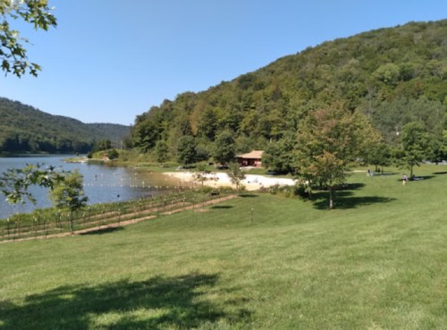 A serene lakeside view with a sandy beach, surrounded by green hills and trees under a clear blue sky.