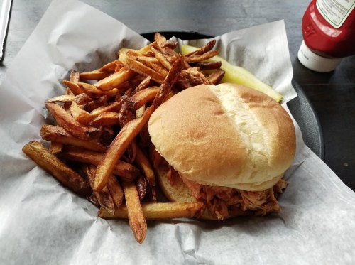 A plate with a pulled pork sandwich and a generous serving of crispy French fries, accompanied by a pickle and ketchup.