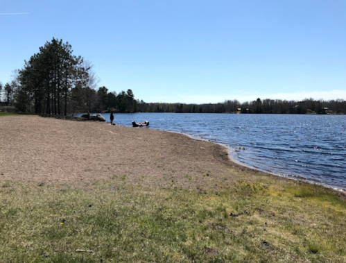 A serene lakeside scene with a sandy beach, trees, and a calm water surface under a clear blue sky.