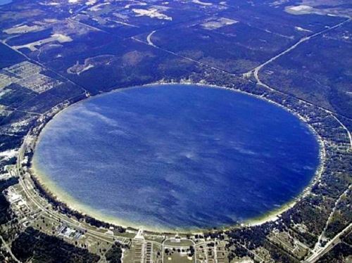 Aerial view of a large circular lake surrounded by forested land and roads.