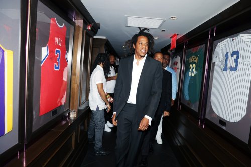 Jay-Z walks through a hallway lined with framed sports jerseys, accompanied by others in formal attire.