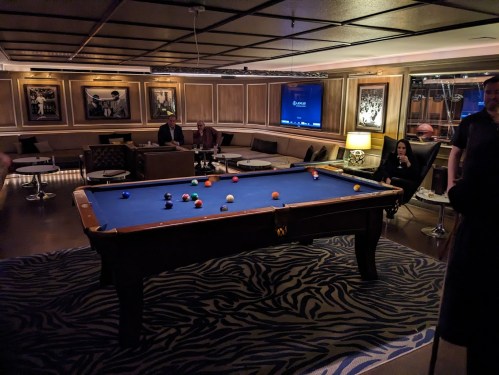 A dimly lit lounge with a blue pool table, surrounded by seating and framed artwork on the walls.