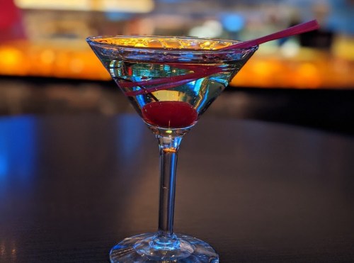A close-up of a colorful cocktail in a martini glass, featuring a cherry and two straws, set against a blurred bar background.