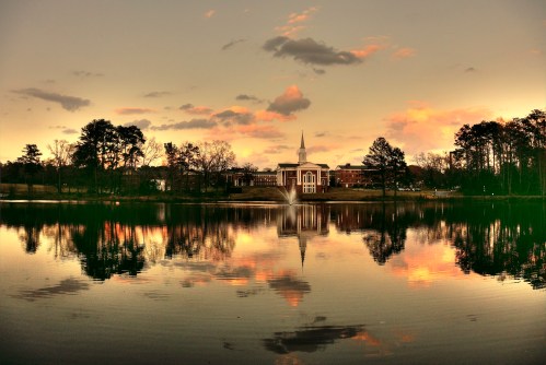 A serene lake reflects a church and trees at sunset, with colorful clouds in the sky.