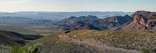 A panoramic view of rugged mountains and valleys under a clear sky, with a winding road in the foreground.