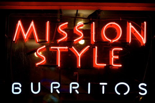 Neon sign reading "MISSION STYLE" in red and "BURRITOS" in blue, glowing against a dark background.