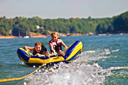 Two children enjoy tubing on a lake, being pulled by a boat, with splashes of water around them and trees in the background.