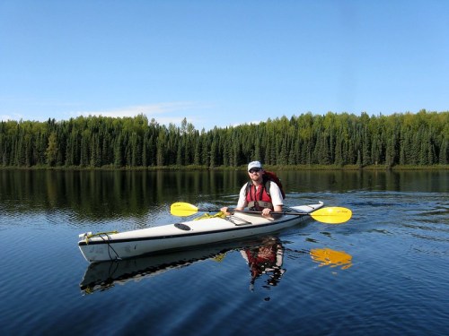 A person in a kayak paddles on a calm lake surrounded by lush green trees under a clear blue sky.