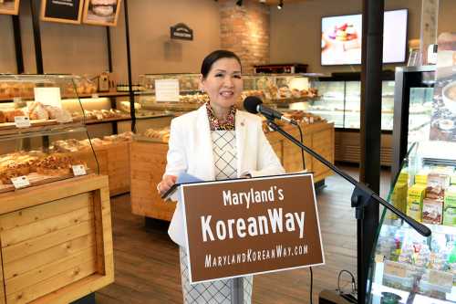 A woman speaks at a podium with "Maryland's KoreanWay" sign in a bakery setting, surrounded by baked goods.