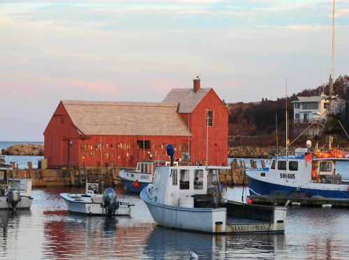 A red barn-like building by the water, with fishing boats docked nearby and a calm sea reflecting the sky.