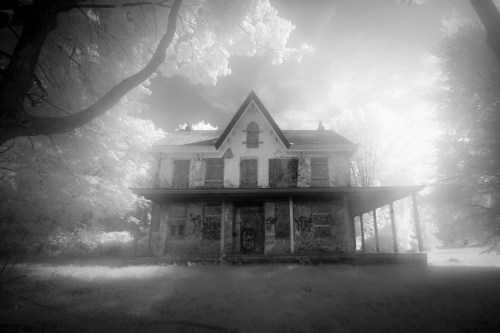 An eerie, abandoned house with boarded windows, surrounded by trees, captured in a misty, black-and-white infrared style.