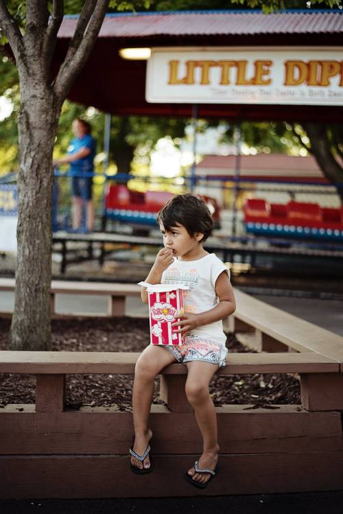 A young child sits on a wooden bench, eating popcorn, with a colorful amusement park in the background.