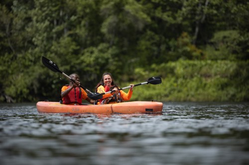 Two people in an orange kayak paddle together on a calm river, surrounded by lush greenery.
