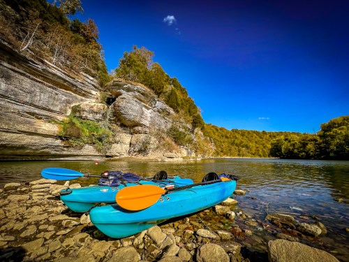 Two blue kayaks with orange paddles rest on a rocky shore beside a calm river, surrounded by autumn foliage and cliffs.