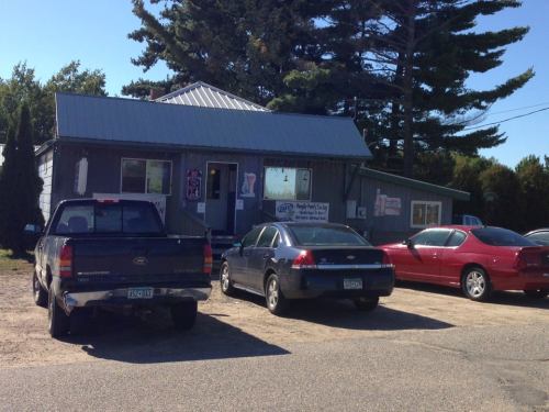 A small gray building with a metal roof, surrounded by parked cars and trees, on a sunny day.