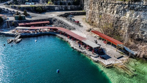 Aerial view of a rocky shoreline with red-roofed structures by a clear blue water body, surrounded by a quarry landscape.