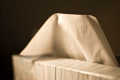 A close-up of a tissue box with a single tissue partially pulled out, set against a softly blurred background.