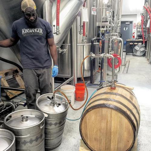 A person in a brewery setting operates equipment near kegs and a wooden barrel.