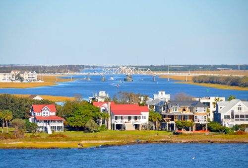 A scenic view of a river with houses along the shore and a bridge in the background under a clear blue sky.
