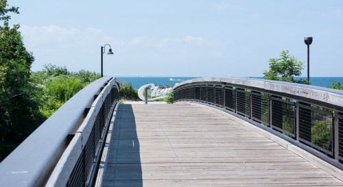 A wooden bridge leads to a scenic view of the ocean, surrounded by greenery and a clear blue sky.