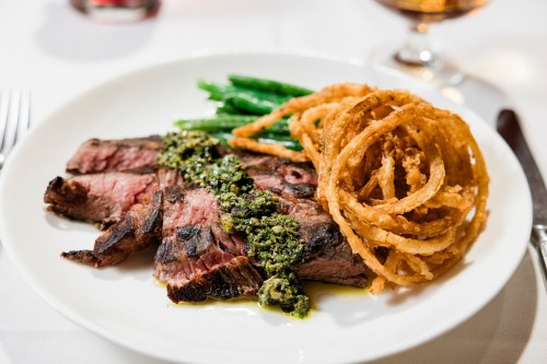 A plate of grilled steak topped with green sauce, served with green beans and crispy onion rings.